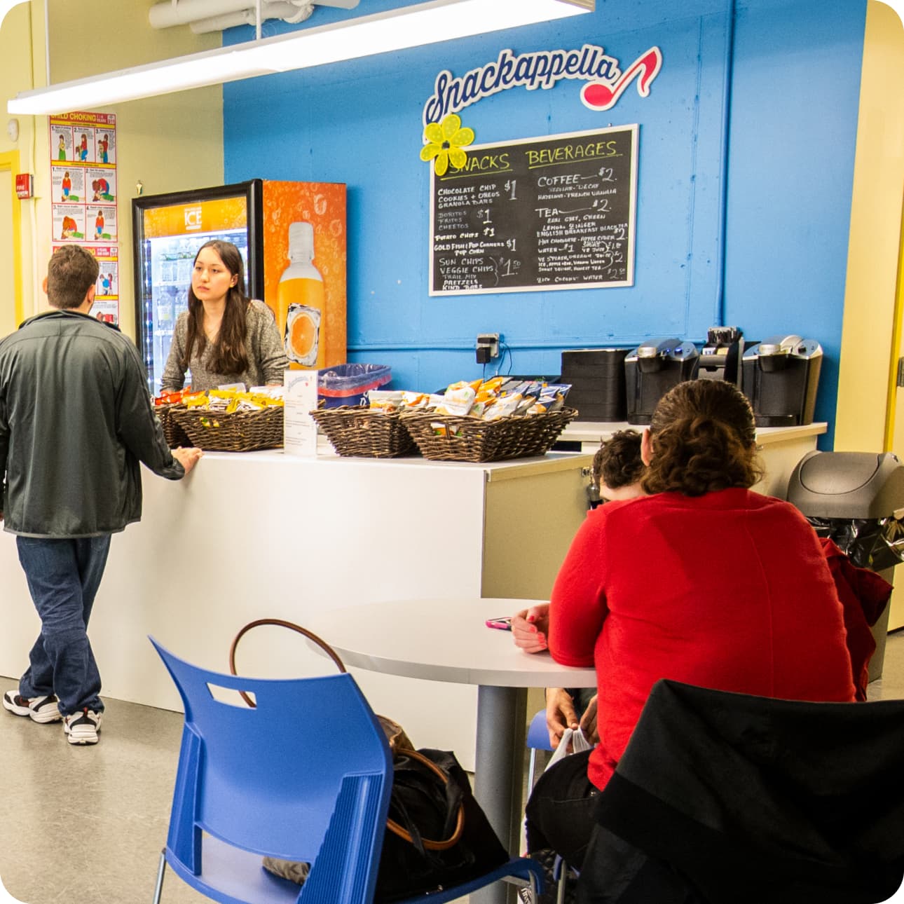 2 friends talk at a table at the Snackappella Cafe as a DMF participant orders a drink. The menu has a musical note and a flower
