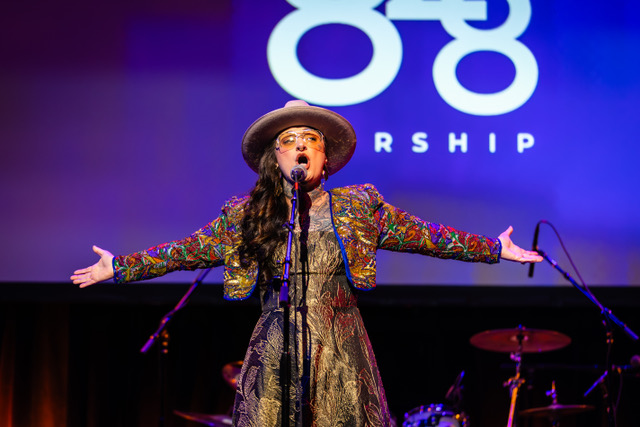 Woman wearing sunglasses and hat singing into a microphone with arms wide open