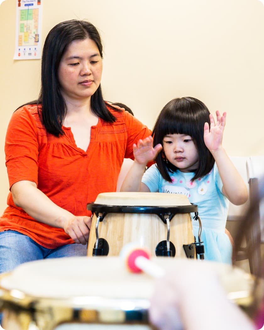 A DMF participant plays a drum with their hands as their parent encourages them