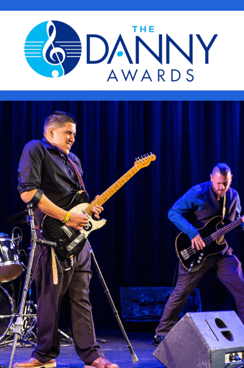 Photo of a Jake Nielsen playing electric guitar on stage with his band. Jake has short hair, and is using crutches. Above him is The Danny Awards logo