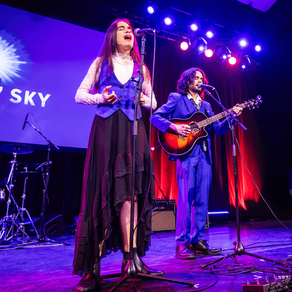 Photo of Jake n' Sky performing at The 2025 Danny Awards. They are a brother and sister duo of light complexion in their teens, wearing blue. The brother is playing guitar and singing behind his sister, who is singing behind a microphone.