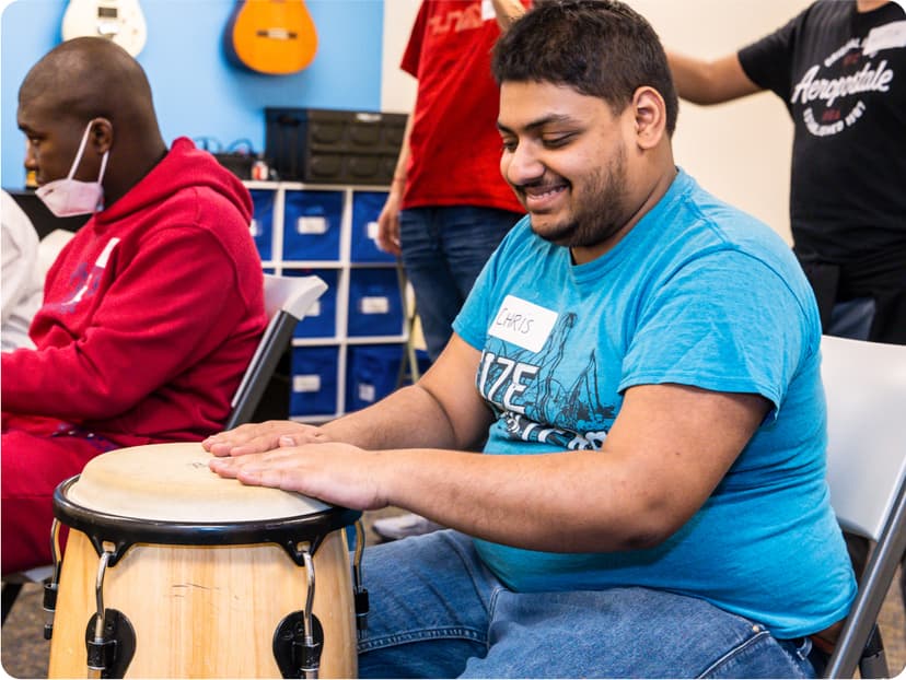 A DMF participant plays a drum with their hands in one of the colorful rooms of the music center. The room is full of people and guitars hang on the wall