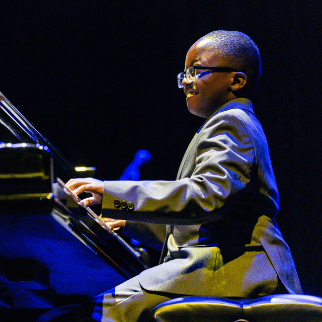 Photo of Jude Kofie performing on a grand piano at The Danny Awards. Jude, an African-American adolescent, is wearing glasses and a grey suit.