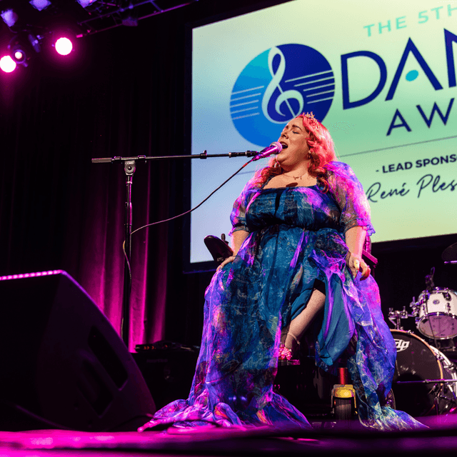 Photo of Marna perfoming at the Danny Awards. She is sitting in a wheelchair and singing into a microphone on the stand. She has blond hair, a light complexion, and is wearing a long purple dress.