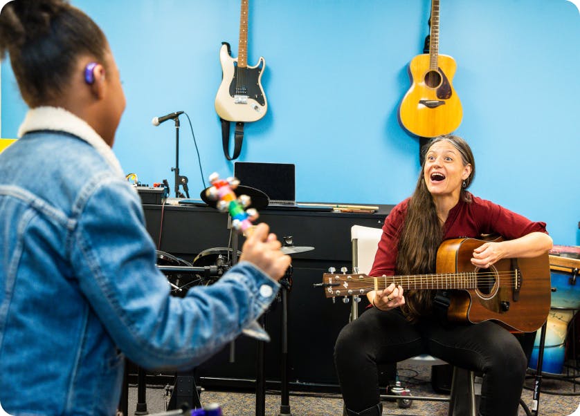 A DMF participant shakes an instrument with jingle bells while DMF Director of Teaching and Learning Juliana plays a guitar and sings