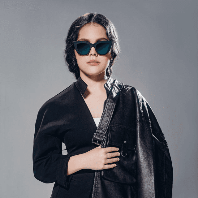 Photo of a young woman wearing dark glasses and a black coat. She has shoulder-length brown hair.