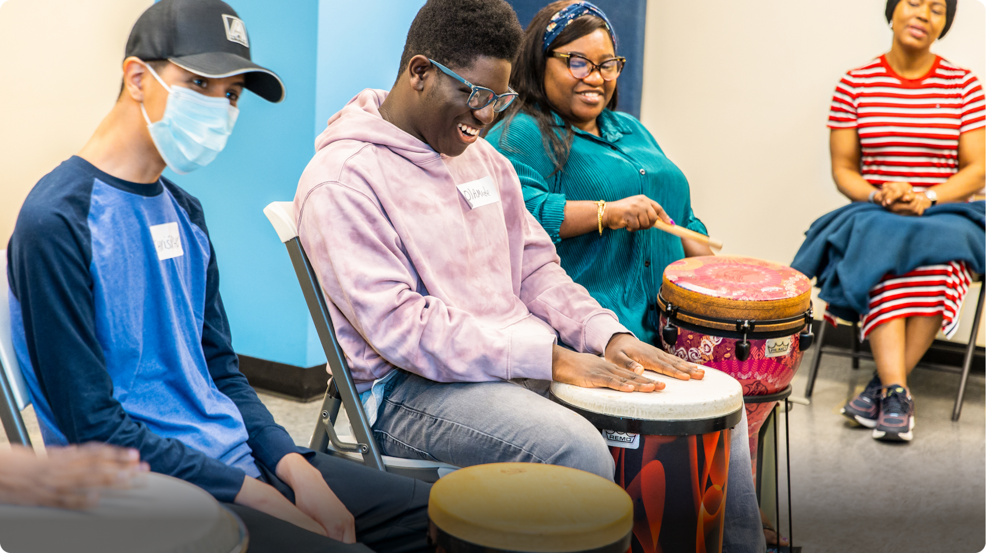 3 DMF participants laugh together and play drums with their hands, while another person supports them in the background