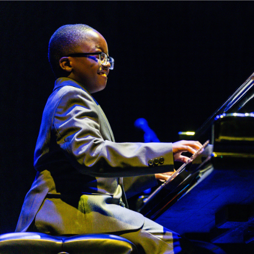 Image of Jude Kofie playing a grand piano. He is a young boy with glasses
