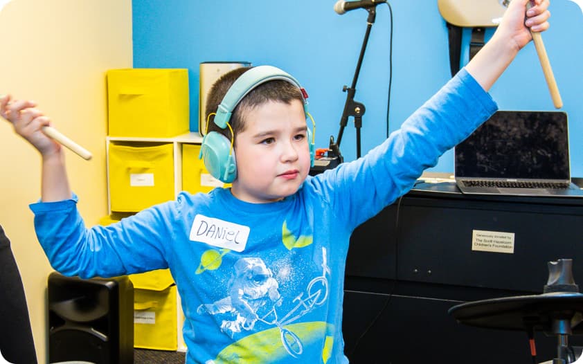 A DMF participant wearing bright blue headphones raises their arms while holding drum sticks