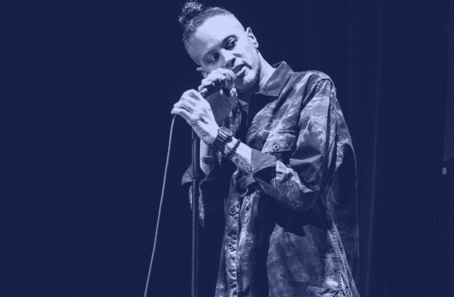 Photo of James Ian standing on stage holding a microphone on a stand. He is wearing a camo-patterened button-down shirt, and has his hair tied up in a bun.