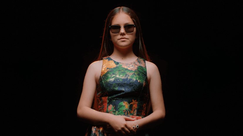 Photo of Iolanta standing in front of a black background. She is a young white woman, wearing dark glasses and a colorful dress.