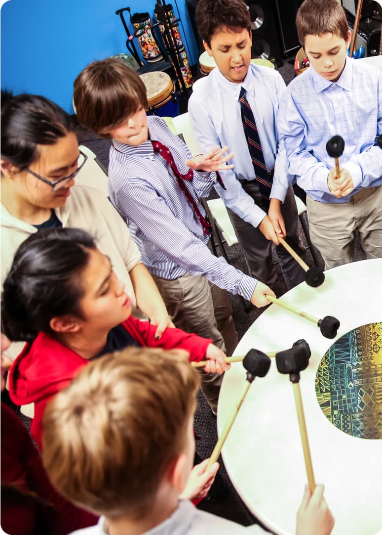 A circle of students play a large drum with drum sticks