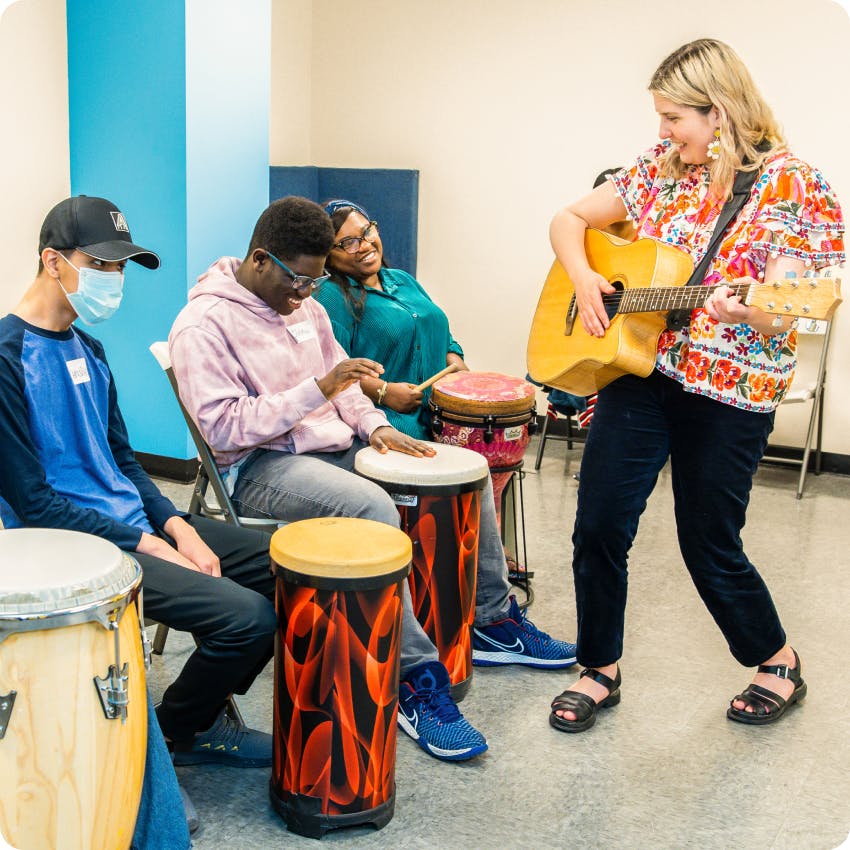 In the music center, DMF Instructor Stefani joyfully plays a guitar and 3 DMF participants play colorful drums