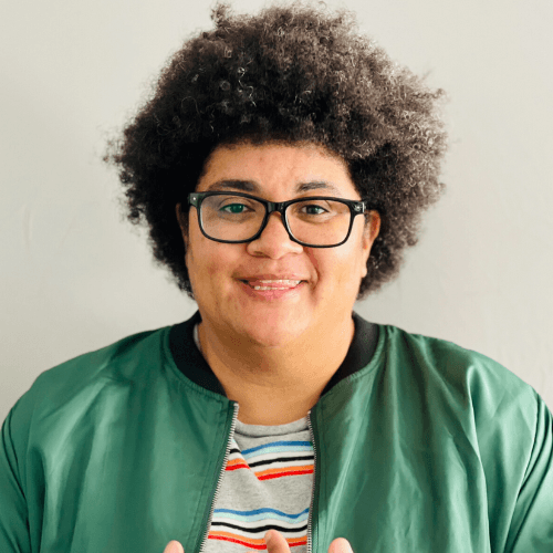 Photo of Jennifer Msumba. A Black woman with an afro in her early thirties, smiling and wearing glasses and a green jacket
