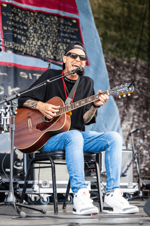 Photo of James Ian singing and playing guitar on stage at Hello Inclusion. He is wearing sunglasses and a backwards baseball cap
